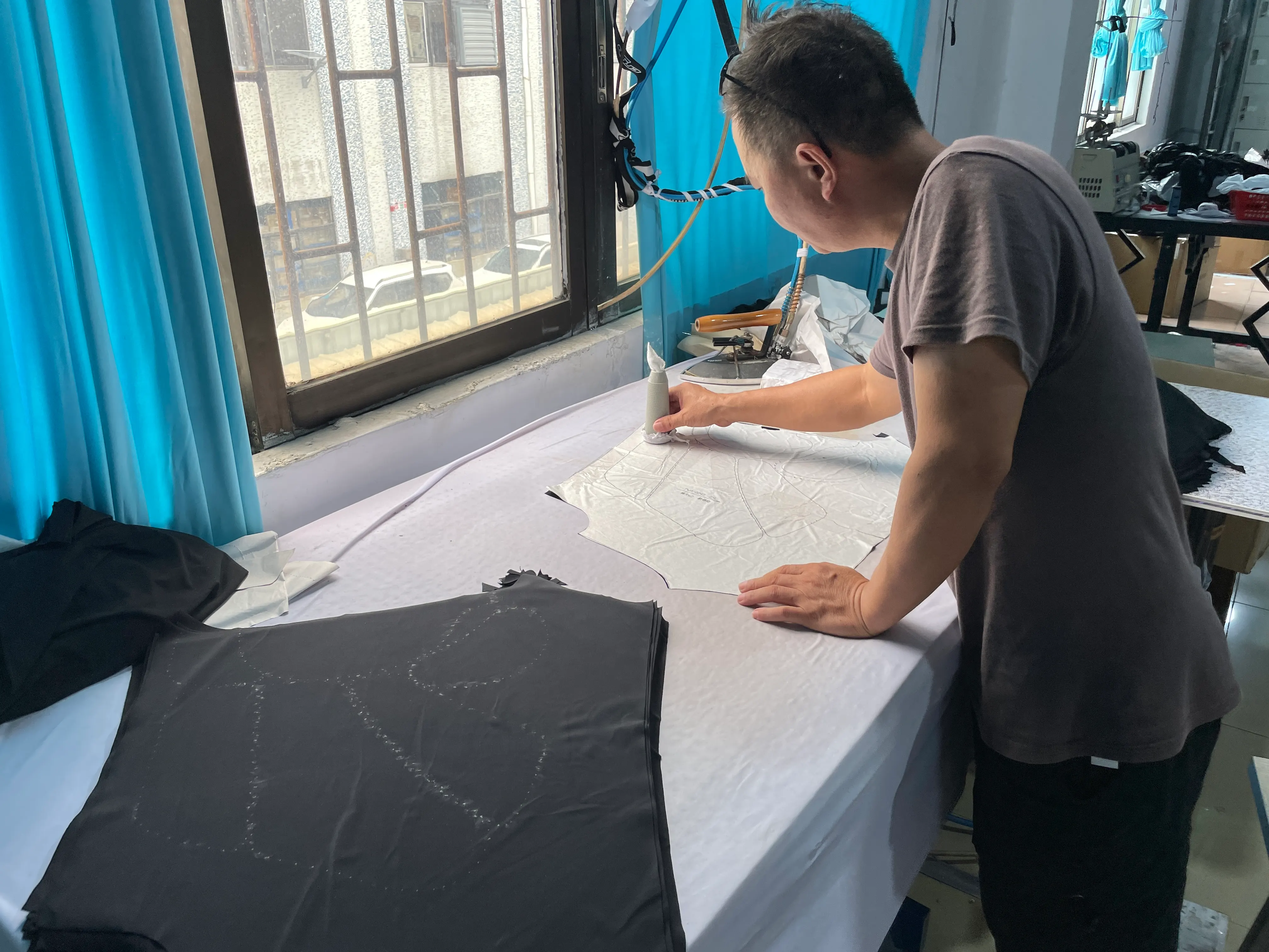 Worker tracing a garment pattern at a worktable beside black fabric pieces.